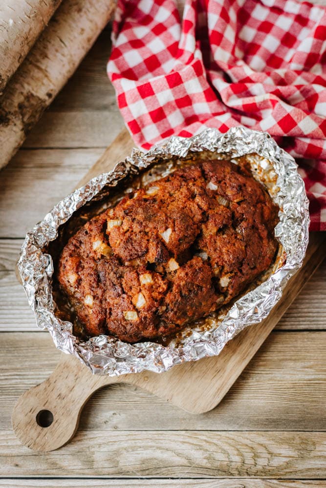 A loaf of bread with visible chunks, baked in aluminum foil like a classic foil packet meatloaf recipe, sits on a wooden cutting board next to a red and white checkered cloth, on a rustic wooden table.
