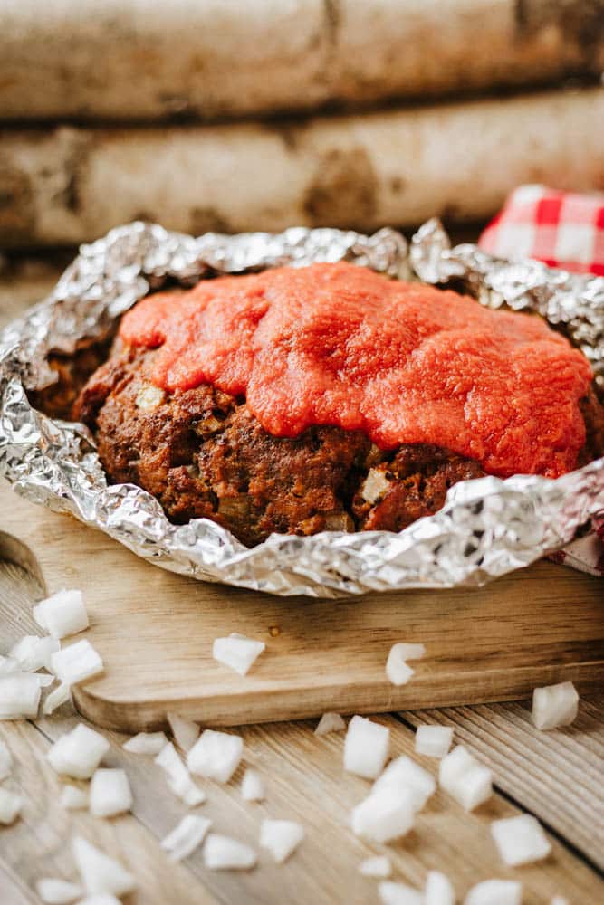 A foil packet meatloaf recipe is showcased here—meatloaf covered in tomato sauce, resting on a wooden board, partially wrapped in aluminum foil with chopped onions nearby. Logs and a red-checkered cloth add rustic charm to the scene.