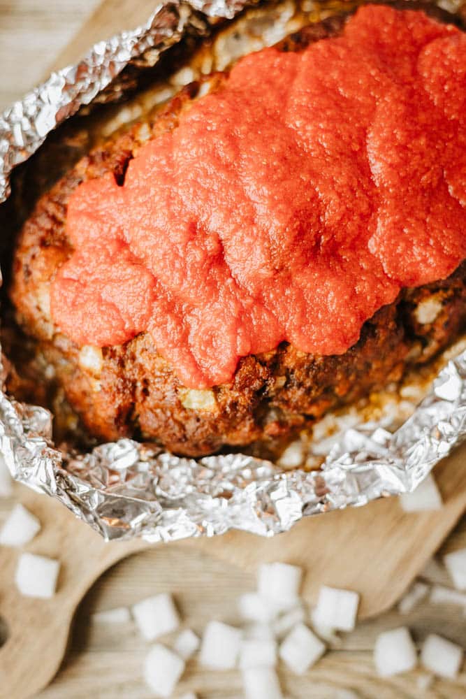 A foil packet meatloaf recipe features a hearty meatloaf covered with red tomato sauce, sitting on aluminum foil atop a wooden board, with small diced onions scattered around.