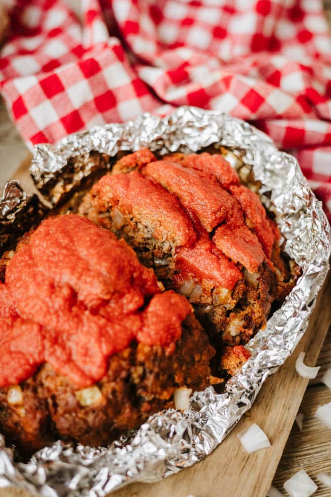 A close-up of a foil packet meatloaf recipe topped with tomato sauce, baked in aluminum foil. Chopped onions are visible in the meatloaf, and a red and white checkered cloth is in the background.