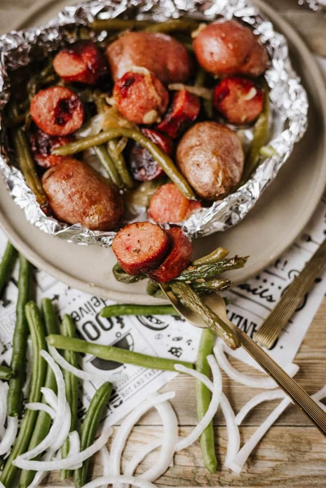 A foil packet sausage and green beans meal, with roasted potatoes, sits on a beige plate. Fresh green beans and sliced onions are scattered nearby, adding a farm-fresh touch to this comforting dish.