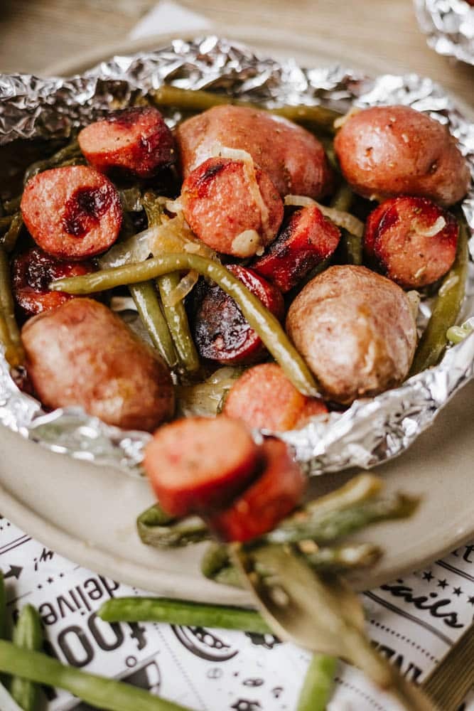 A foil packet sausage and green beans meal featuring cooked small potatoes, sliced sausage, and green beans, all perfectly seasoned and roasted, served on a plate.