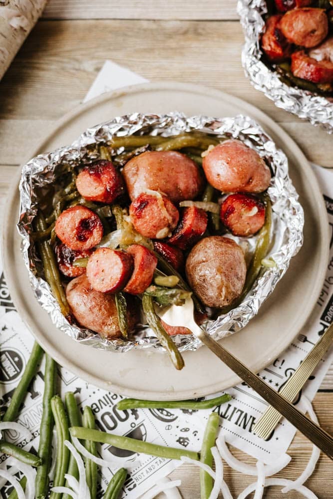 Foil packet sausage and green beans with baby potatoes are served on a beige plate, surrounded by fresh green beans and onion slices on a wooden table.