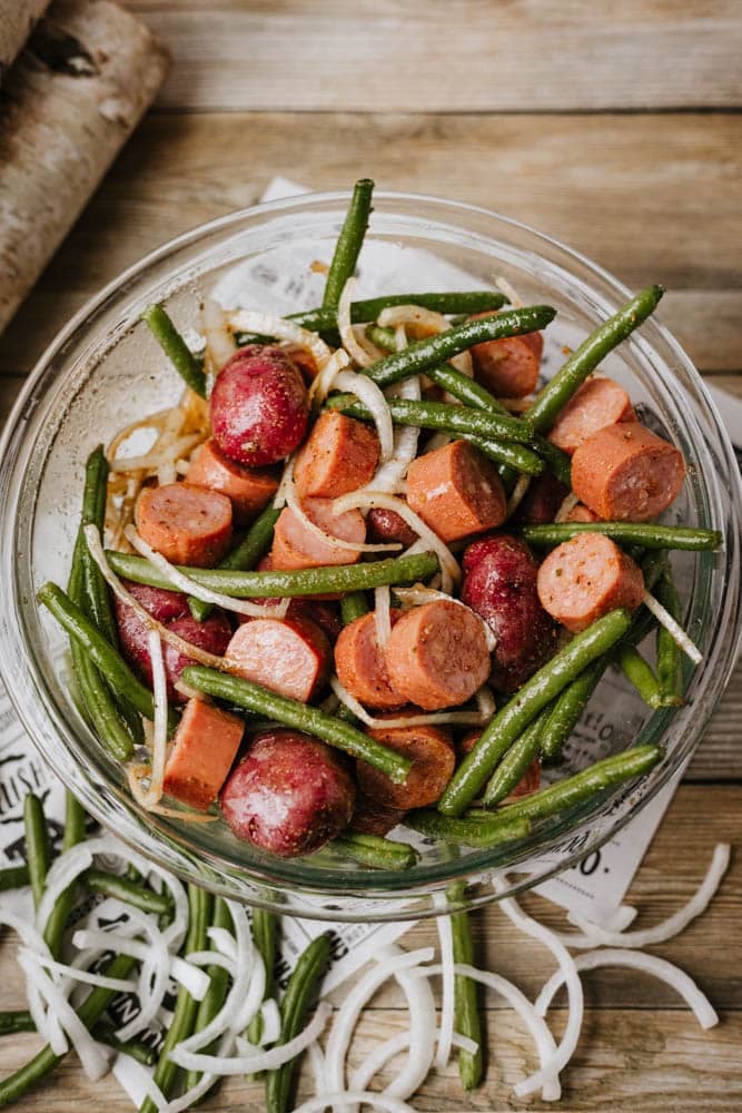 A glass bowl filled with sliced sausage, green beans, red potatoes, and onion slices—just like a classic foil packet sausage and green beans recipe—sits on a wooden surface with raw onion rings scattered beside it.