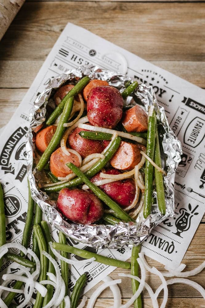 A foil packet filled with sausage, red potatoes, green beans, sliced carrots, and onion rings, seasoned and ready for cooking, sits on printed parchment atop a wooden surface.