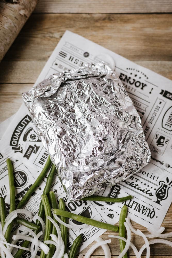 A foil packet sausage and green beans meal sits on decorative printed paper, with sliced green beans and raw onion rings scattered nearby on a wooden surface.