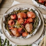 A foil packet sausage and green beans meal, featuring sliced sausages, whole baby potatoes, and green beans, is served on a beige plate. Nearby, uncooked green beans and sliced onions are scattered on a wooden surface.