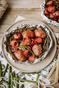 A foil packet sausage and green beans meal, featuring sliced sausages, whole baby potatoes, and green beans, is served on a beige plate. Nearby, uncooked green beans and sliced onions are scattered on a wooden surface.