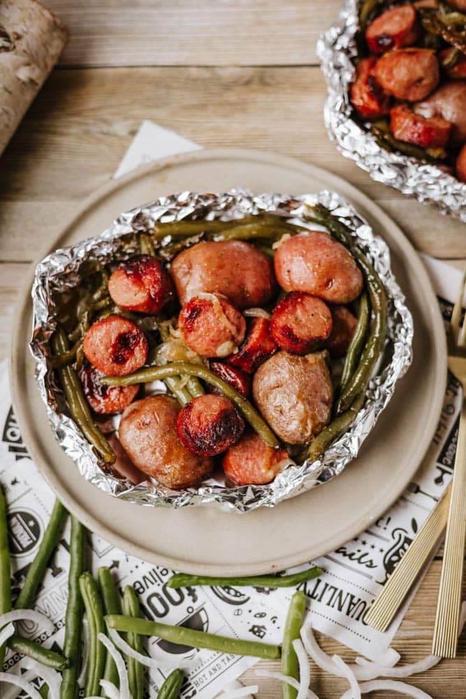 A foil packet sausage and green beans meal, featuring sliced sausages, whole baby potatoes, and green beans, is served on a beige plate. Nearby, uncooked green beans and sliced onions are scattered on a wooden surface.