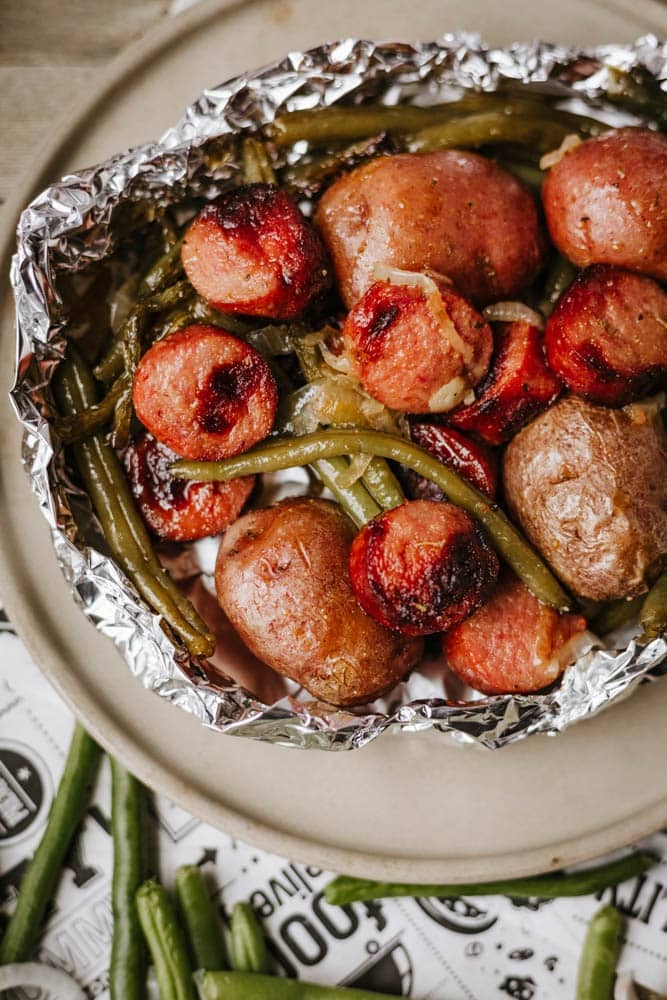A foil packet of sausage and green beans with roasted red potatoes sits on a plate, while extra green beans rest on the table beside it.