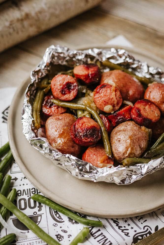 A foil packet sausage and green beans meal with roasted red potatoes sits on a beige plate, accented by fresh green beans scattered nearby.