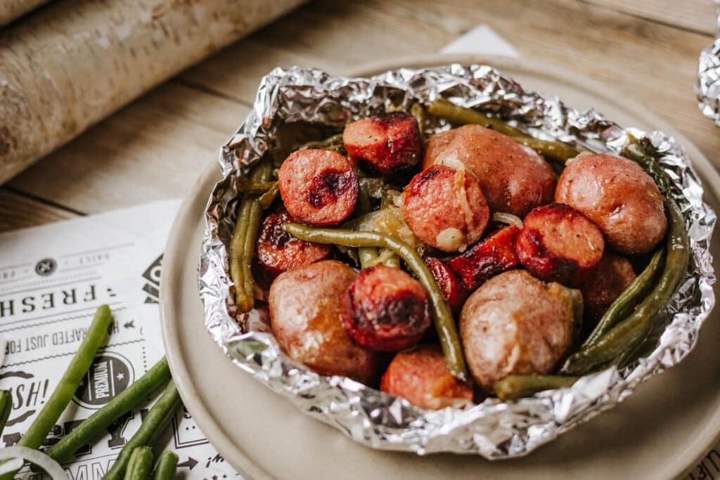 A foil packet sausage and green beans dish with roasted potatoes sits on a beige plate, surrounded by extra green beans and part of a newspaper in the background.