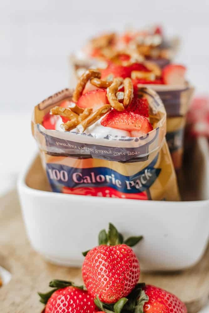 A pack of 100-calorie chocolate pudding topped with whipped cream, fresh strawberry slices, and pretzel twists sits in a white dish. Additional whole strawberries are placed in front of the container.