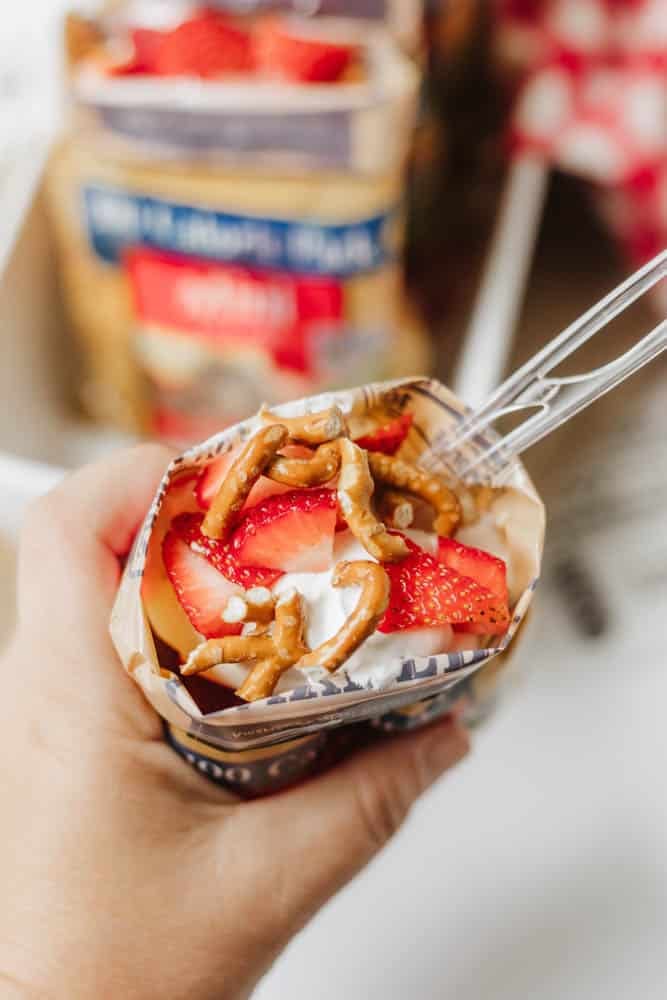 A hand holds an open chip bag filled with whipped topping, sliced strawberries, and pretzel pieces, with a plastic fork inserted.