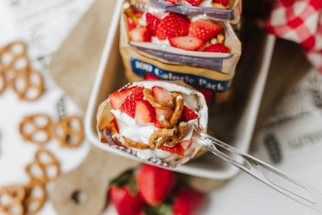 A spoon scoops a snack mix of yogurt, sliced strawberries, and pretzels from an open bag labeled 100 Calorie Pack. More snacks and fresh strawberries are nearby.