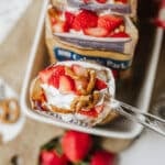 A spoon holding a dessert made of pretzels, whipped cream, and sliced strawberries, with more servings in pretzel snack bags arranged on a tray below. Fresh strawberries and pretzels are scattered nearby.