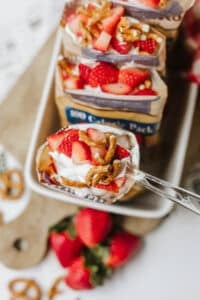 A spoon holding a dessert made of pretzels, whipped cream, and sliced strawberries, with more servings in pretzel snack bags arranged on a tray below. Fresh strawberries and pretzels are scattered nearby.