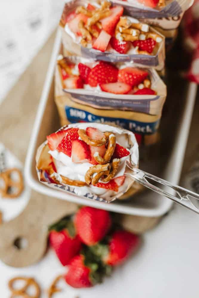 A spoon holding a dessert made of pretzels, whipped cream, and sliced strawberries, with more servings in pretzel snack bags arranged on a tray below. Fresh strawberries and pretzels are scattered nearby.