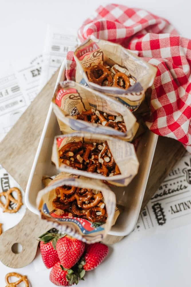 Four paper snack bags filled with pretzels are arranged in a white tray on a wooden board. A red checkered cloth and three strawberries are nearby, with some pretzels scattered on the surface.
