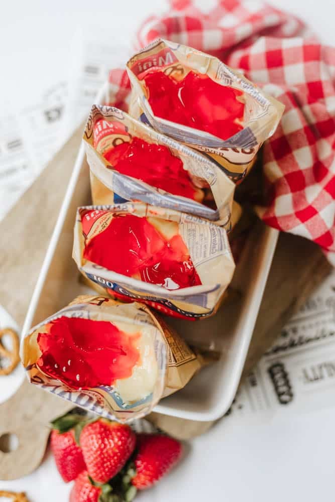 Four open vanilla pudding snack bags topped with bright red strawberry gelatin are arranged in a white dish, with a checkered red cloth and fresh strawberries nearby.
