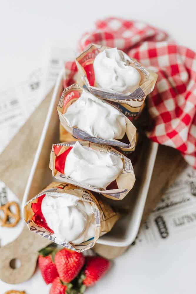 Four open snack-size chip bags filled with whipped cream are arranged in a white tray, with a red-and-white checkered cloth and fresh strawberries nearby on a wooden board.