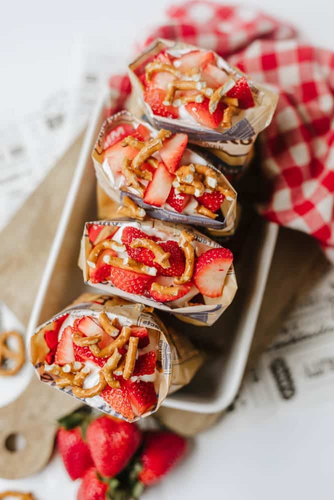 Four snack-sized paper bags filled with yogurt, sliced strawberries, and pretzel pieces are arranged on a tray, with fresh strawberries and a red checkered cloth in the background.