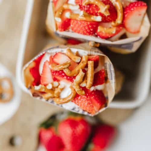 Overhead view of a snack bag opened and filled with whipped cream, sliced strawberries, and pretzels, placed on a tray with fresh strawberries and pretzels nearby.