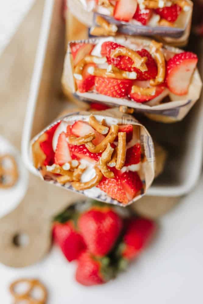 Overhead view of a snack bag opened and filled with whipped cream, sliced strawberries, and pretzels, placed on a tray with fresh strawberries and pretzels nearby.